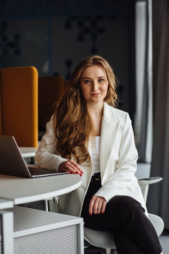 Beautiful stylish young business lady working on the camera at the desk. Portrait of a smiling woman in a business suit. A girl with long hair and a white jacket is sitting at a desk.