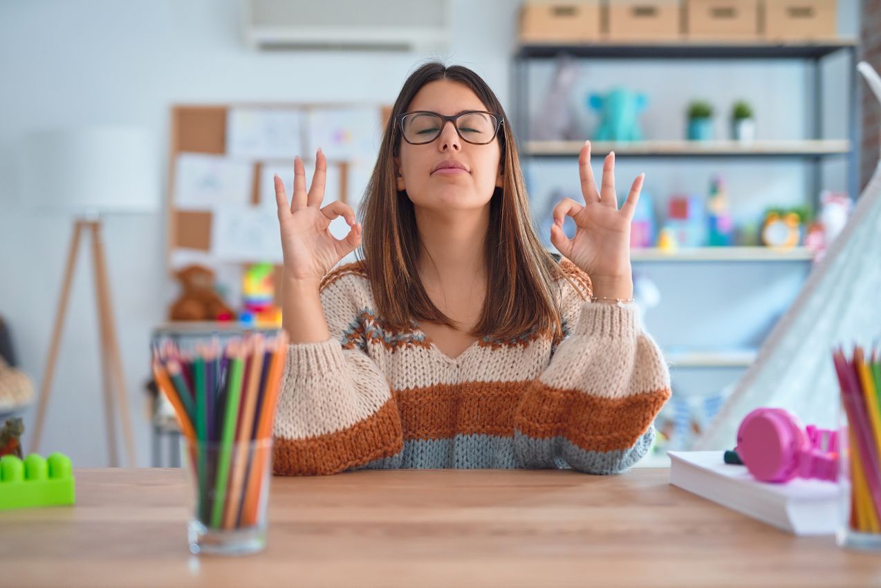 Young beautiful teacher woman wearing sweater and glasses sitting on desk at kindergarten relaxed and smiling with eyes closed doing meditation gesture with fingers. Yoga concept. Enseignante détendue dans une classe bien aérée grâce à une VMC performante